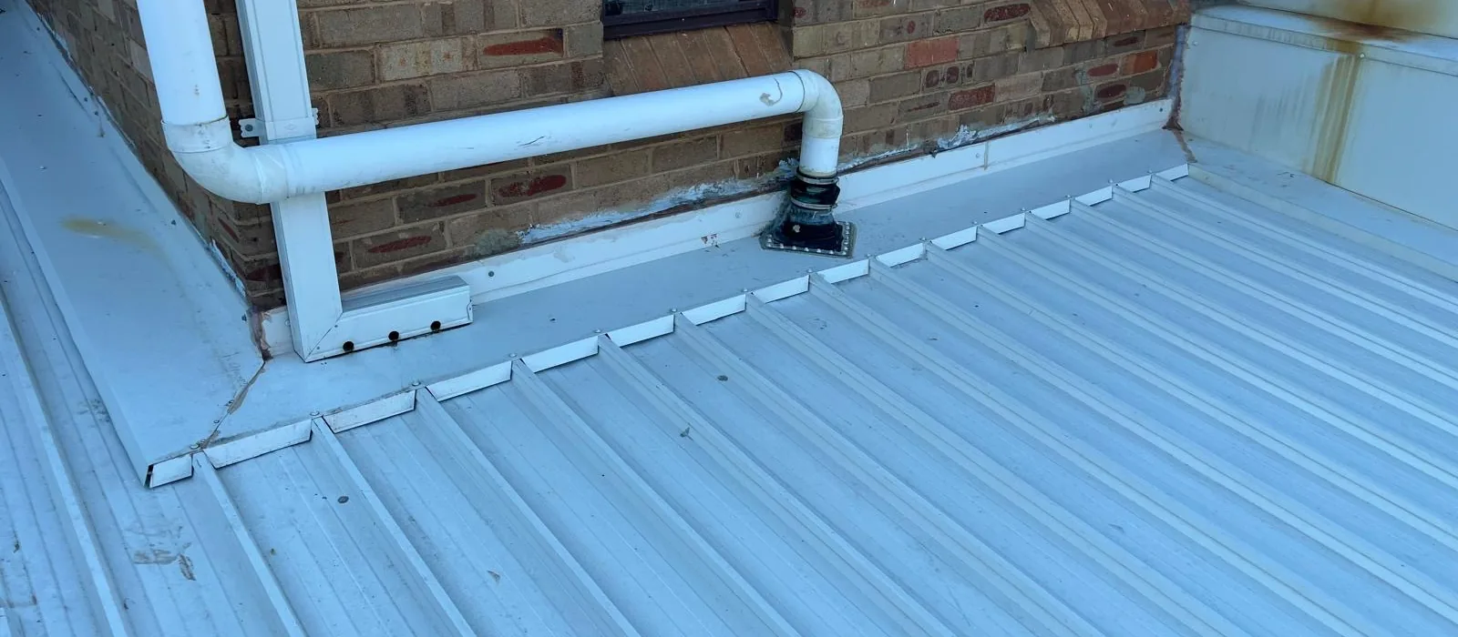 Low-pitch metal deck roof meeting brick, with a sealed vent penetration